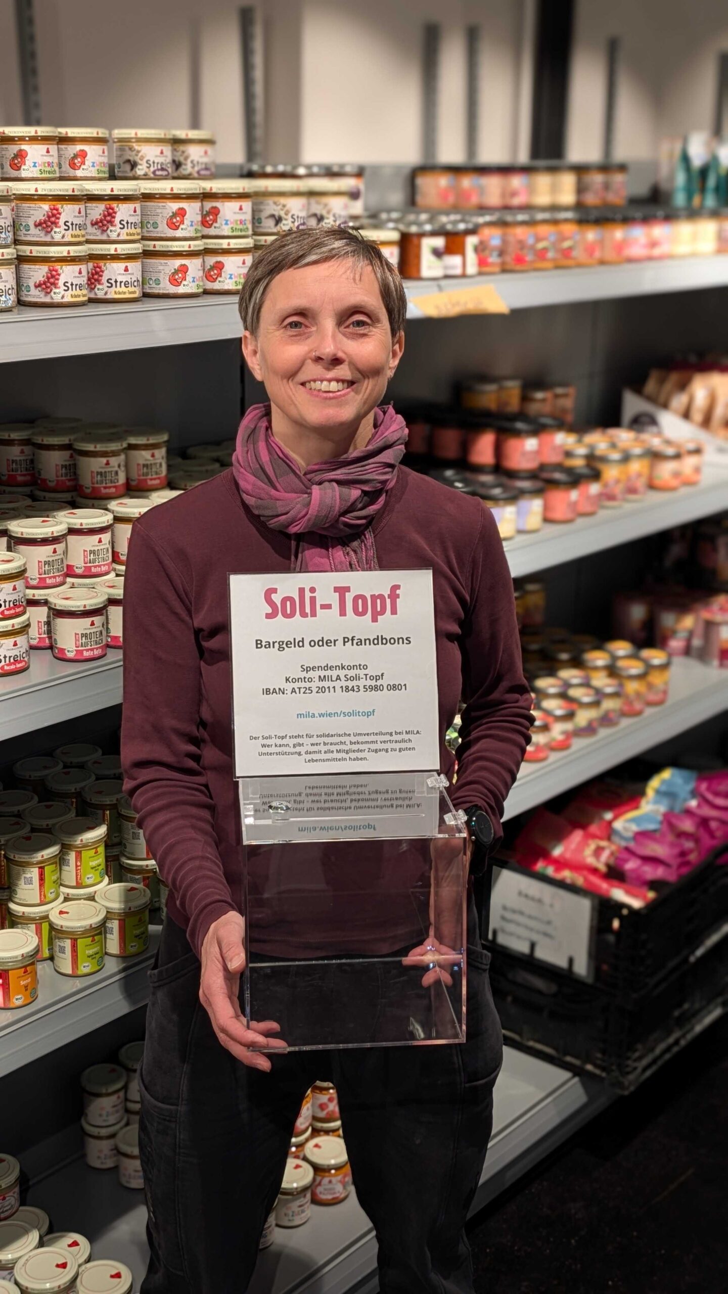 Soli-Topf MILA Mitglied hält Glasbox vor gefülltem regal