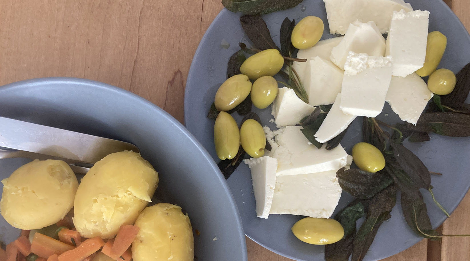 Paneer (indischer Frischkäse) mit Oliven und gebratenem Salbei auf einem Teller angerichtet, daneben ein teller mit gekochten Erdäpfeln und Karottengemüse.